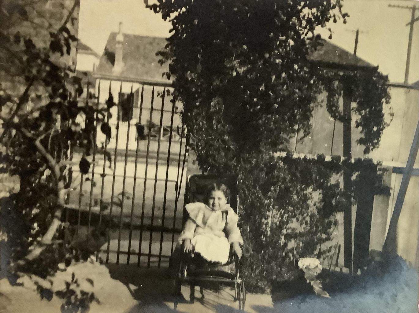 A yellowed black and white photo of a girl in a frilly dress sitting in a wheelchair. Sheehan posed in front of a large house with vines growing decoratively over the gate