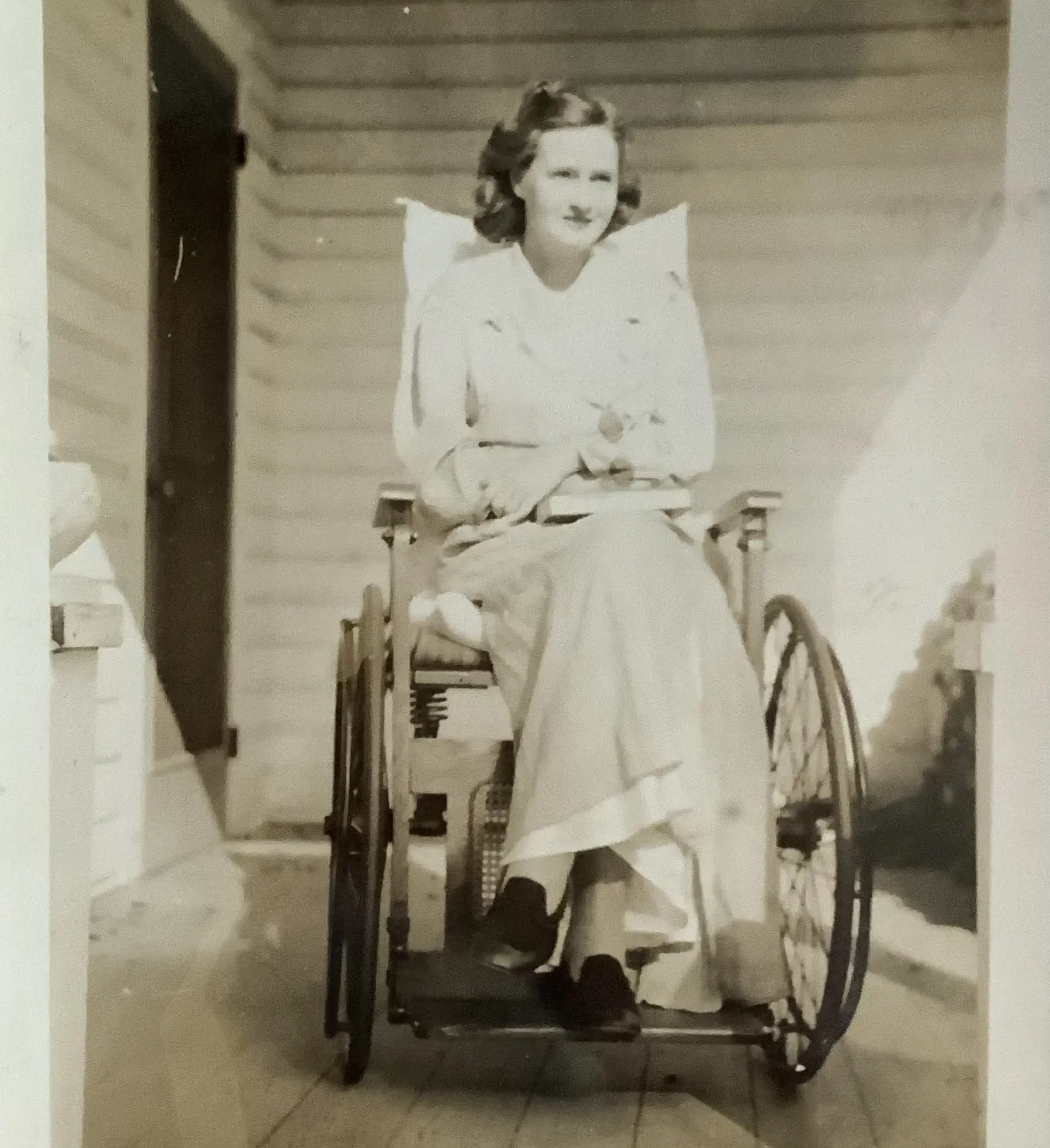 A yellowed black and white photo of a girl in a frilly dress sitting in a wheelchair. Sheehan posed in front of a large house with vines growing decoratively over the gate
