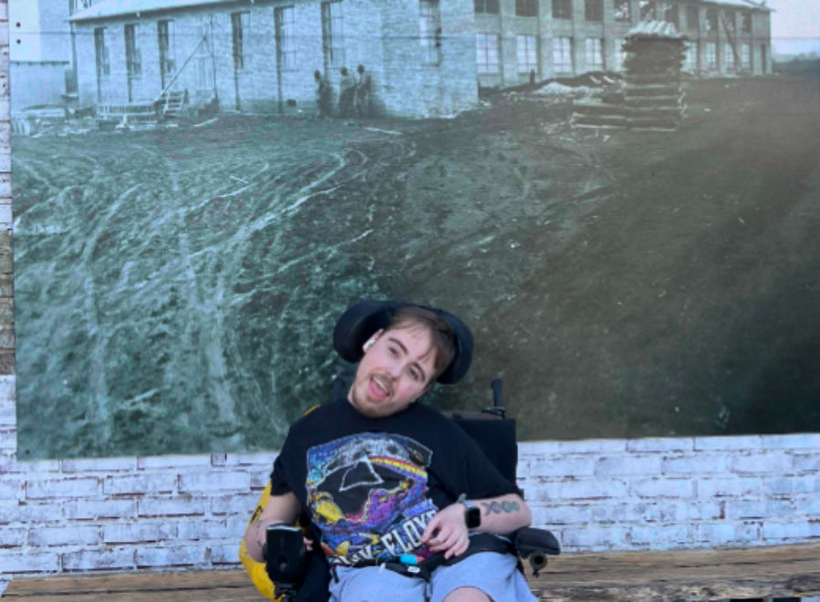 A young man in a powered wheelchair poses in front of a mural on a brick wall. He is white with short brown hair and a beard. He is wearing a Pink Floyd shirt and has a tattoo of DNA on his left arm.