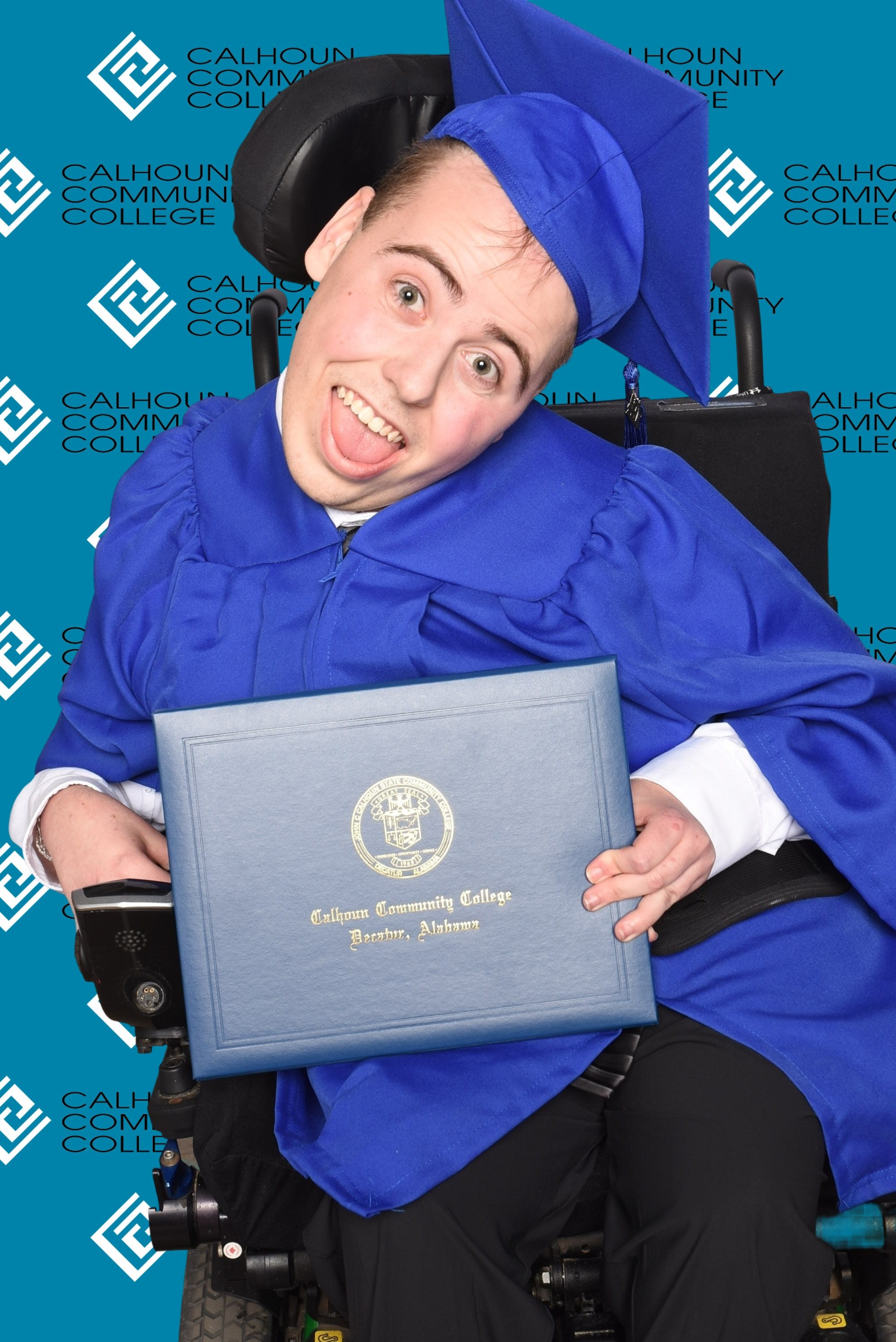 A young man in a powered wheelchair poses in his cap and gown with his college diploma.