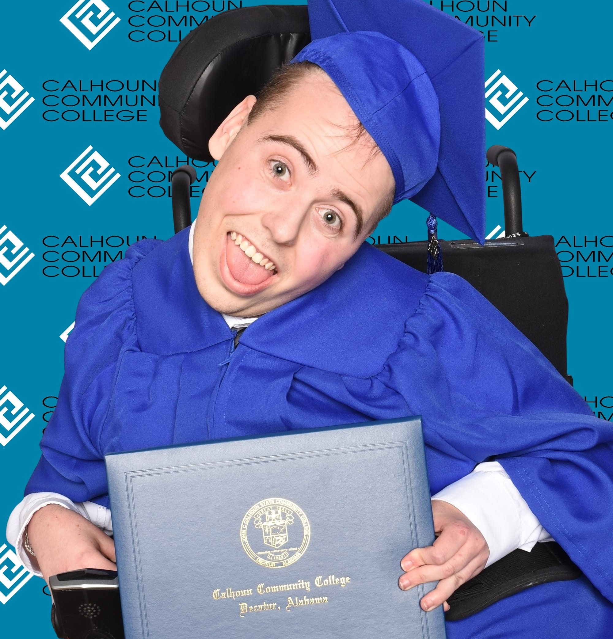 A young man in a powered wheelchair poses in his cap and gown with his college diploma.